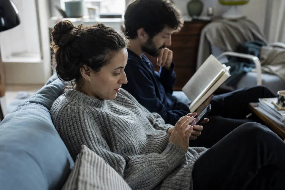 Woman typing a question on her phone on the sofa while her husband reads a book beside her