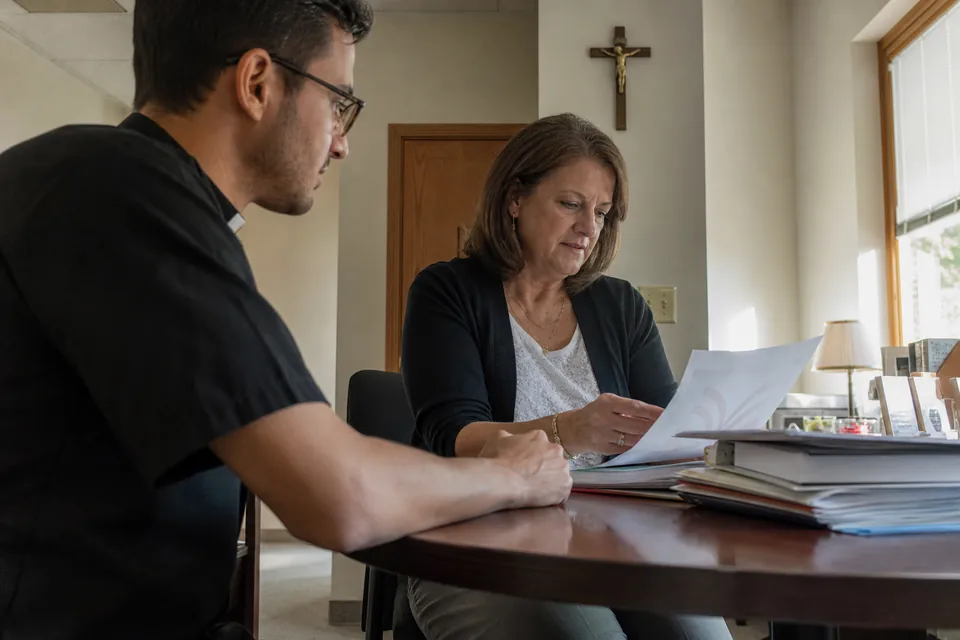 Parish marriage preparation coordinator at an office desk reviewing a printed couple report with a deacon