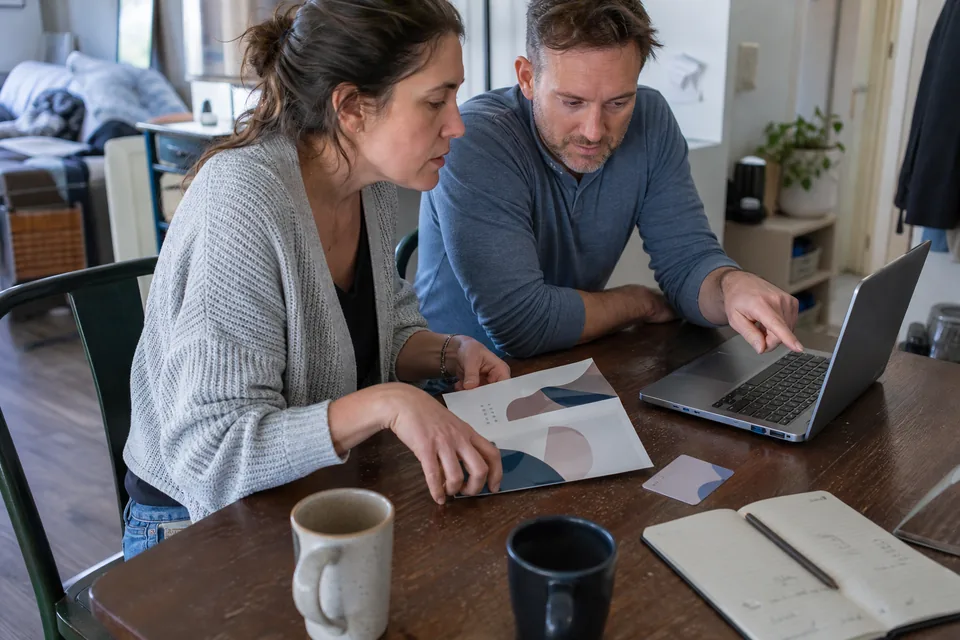 Married couple at a dining table comparing a parish course brochure with an online program on a laptop