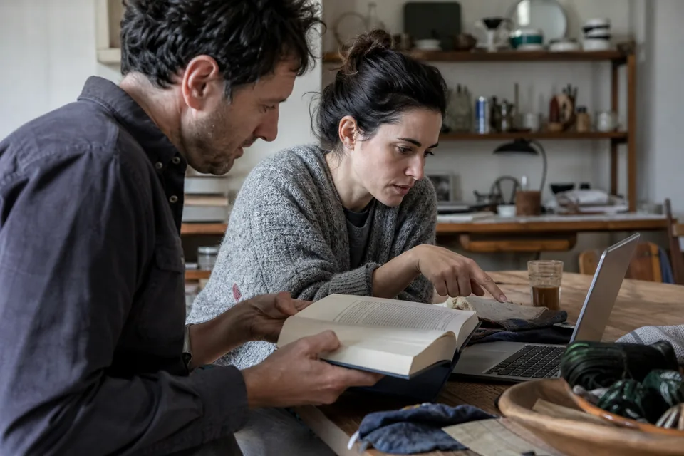 Husband holding open a thick book while his wife compares a paragraph with the laptop beside them