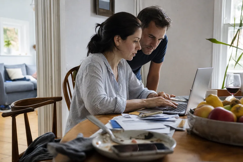 Wife typing a question into a laptop at the dining table while her husband reads over her shoulder