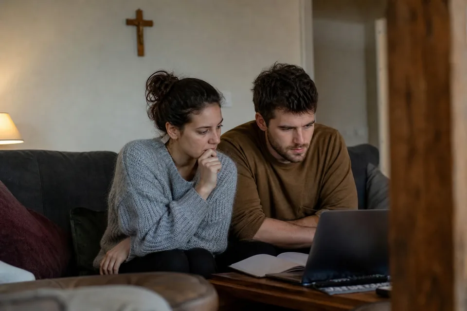 Engaged couple in their late twenties sitting on a living room sofa with a laptop and an open notebook, preparing together in the evening