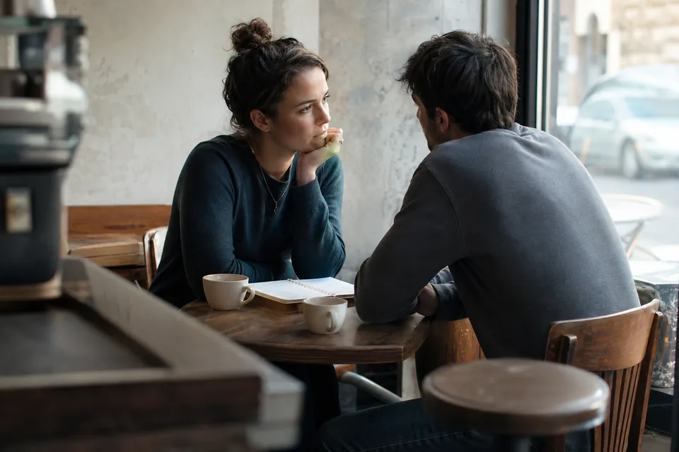 Engaged couple in their twenties sitting at a cafe table with two coffees and a small notebook, talking quietly