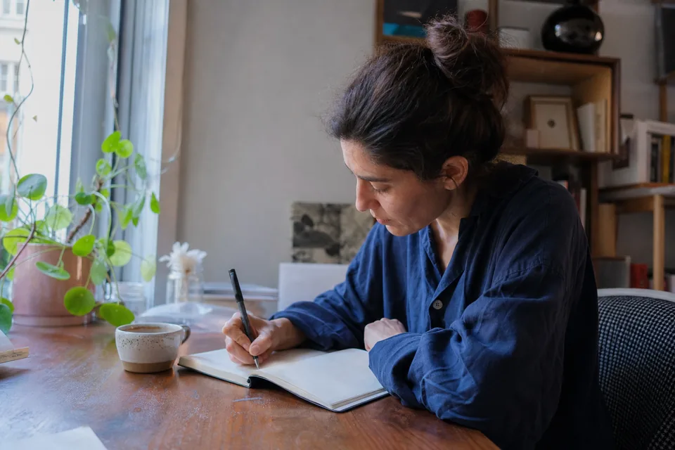 Woman writing in a notebook at a wooden table at home, a cup of tea beside her